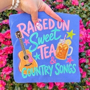 May include: A blue t-shirt with the text "RAISED ON Sweet TEA & COUNTRY SONGS" in pink and teal lettering. The shirt features illustrations of a guitar and a glass of iced tea with a lemon slice.