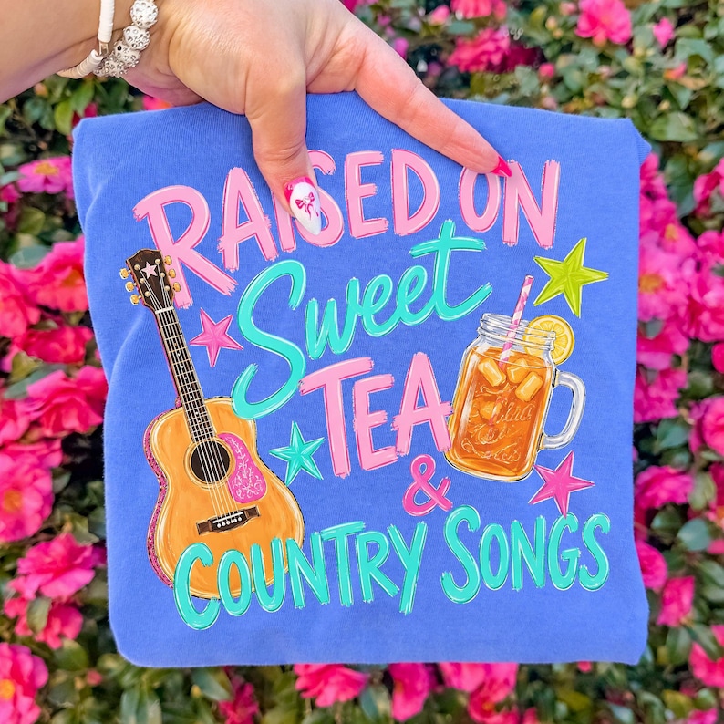 May include: A blue t-shirt with the text "RAISED ON Sweet TEA & COUNTRY SONGS" in pink and teal lettering. The shirt features illustrations of a guitar and a glass of iced tea with a lemon slice.