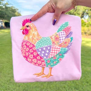 May include: A light pink t-shirt featuring a colorful graphic of a chicken. The chicken is designed with a patchwork pattern consisting of various sections in shades of pink, teal, and purple, decorated with floral, gingham, and star prints.