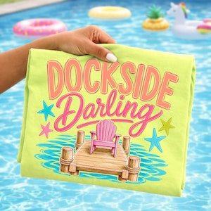 May include: A lime green t-shirt featuring a graphic print with the text DOCKSIDE Darling in peach and pink letters. The design includes a wooden dock, a pink Adirondack chair, and colorful starfish icons set against blue water ripples.