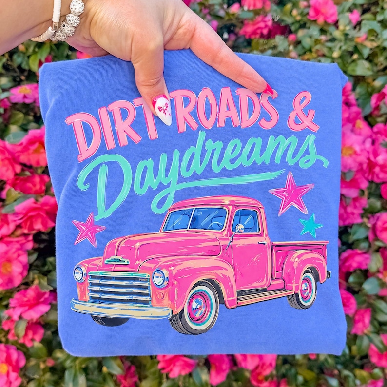 May include: A periwinkle blue t-shirt featuring a graphic of a bright pink vintage pickup truck. Above the truck, the text reads "DIRT ROADS & Daydreams" in pink and teal. The design includes pink and teal star accents surrounding the truck.