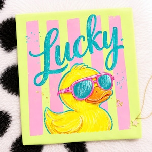 May include: A square canvas art piece featuring a yellow duck wearing pink sunglasses. The word "Lucky" is written in teal script above the duck. The background has vertical pink and light green stripes.