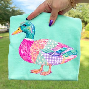 May include: A mint green t-shirt featuring a colorful mallard duck illustration. The duck has a bright blue head, an orange beak, and a body decorated with patchwork patterns including pink leopard print, white stars on teal, and purple gingham checks.