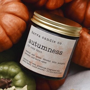 Może przedstawiać: Słoik ze świecą ze złotą pokrywką, z napisem "Autumness" firmy Barva Candle Co. Świeca otoczona jest pomarańczowymi dyniami i zielonymi tykwami. Etykieta wymienia zapach orzecha włoskiego, pekanu, karmelu i ciepłych przypraw.