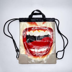 Può includere: Zaino beige e nero con una grande grafica di una bocca aperta con labbra rosse. Lo zaino ha cordini neri e un manico nero. Gli angoli inferiori sono di un colore beige più chiaro.
