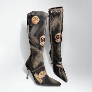 May include: A pair of knee-high denim boots in a faded charcoal wash featuring metallic stiletto heels, silver star studs, and various patches including the text DTS, 66, and el dantes. The boots are adorned with decorative zippers and stitching.