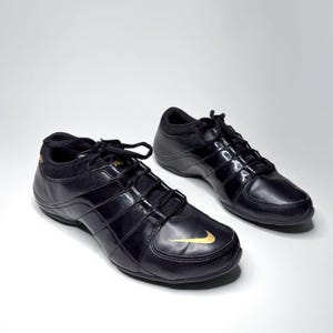 May include: A pair of black athletic sneakers featuring a glossy finish, black laces, and a gold Nike swoosh logo on the toe area. The shoes have a low-top silhouette with a padded collar and a sleek, streamlined design for performance and daily wear.