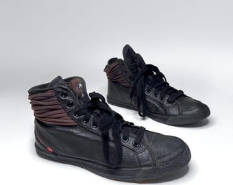 May include: Black high-top sneakers with brown accents. The sneakers feature black laces, a rubber sole, and a layered brown fabric detail around the ankle. The Diesel Industry logo is visible on the side.