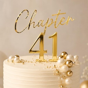May include: A round white frosted cake on a gold stand, decorated with white and gold spheres. A gold acrylic cake topper sits on top, featuring the word Chapter in cursive script above the large numerals 41 in a bold, sans-serif font.