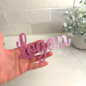 May include: A pink glittery cursive "Logan" name cutout. The name is held in a hand, with a white and gray background. The name is a decorative item, possibly for a party or room decor.