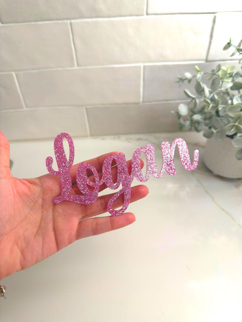 May include: A pink glittery cursive "Logan" name cutout. The name is held in a hand, with a white and gray background. The name is a decorative item, possibly for a party or event.