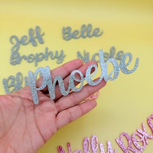 May include: Silver glitter cursive name cutouts, including "Phoebe", "Jeff", and "Belle", are displayed against a yellow background. Other names in pink glitter are also visible. These decorative name cutouts are ideal for personalized crafts.