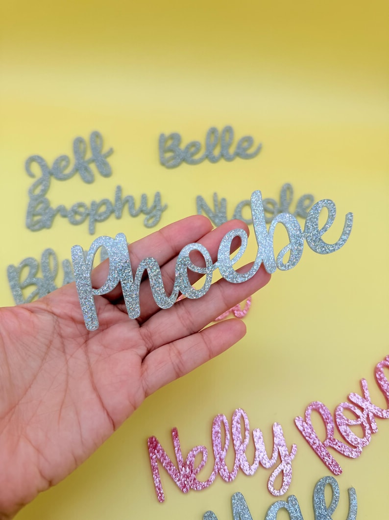 May include: Silver glitter cursive name cutouts, including "Phoebe", "Jeff", and "Belle", are displayed against a yellow background. Other names in pink glitter are also visible. These decorative name cutouts are ideal for personalized crafts.