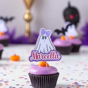 Könnte beinhalten: Ein Halloween-Cupcake mit lila Zuckerguss und einem geisterförmigen Aufsatz mit der Aufschrift "Marcella". Der Cupcake ist mit einer orangefarbenen Süßigkeit verziert und steht auf einer weißen Oberfläche, die mit Konfetti übersät ist.