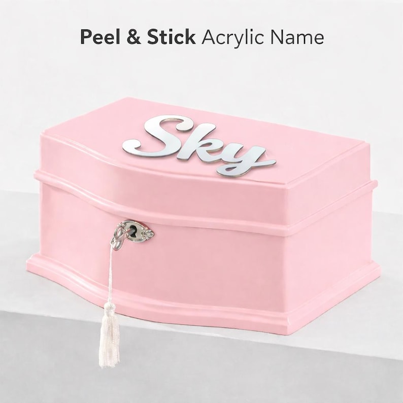 May include: A light pink jewelry box with a curved lid featuring the silver acrylic name Sky in cursive script. The box includes a silver metal lock mechanism with a hanging white tassel. The text Peel & Stick Acrylic Name is visible.