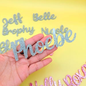 May include: Glittery name cutouts in silver and pink, with names like Phoebe, Nelly, and Roxie. The names are in a cursive font, perfect for party decorations or personalized gifts. The background is a bright yellow.