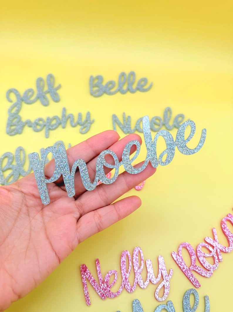 May include: Glittery name cutouts in silver and pink, with names like Phoebe, Nelly, and Roxie. The names are in a cursive font, perfect for party decorations or personalized gifts. The background is a bright yellow.