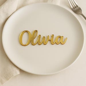 May include: A gold-colored acrylic name tag featuring the name Olivia in a cursive script font. The name tag is placed on a plain white ceramic plate, with a silver-colored metal fork visible at the top right corner of the plate.