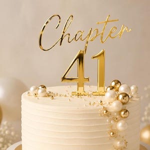 May include: A gold-coloured cake topper featuring the word Chapter in cursive script above the number 41 in a bold serif font. The topper is placed on a white frosted cake decorated with a cluster of white and gold spheres of varying sizes.