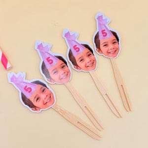 May include: Four wooden skewers with a smiling child's face and a pink party hat with the number 5 printed on it. The skewers are arranged on a light yellow surface, suggesting a birthday theme.