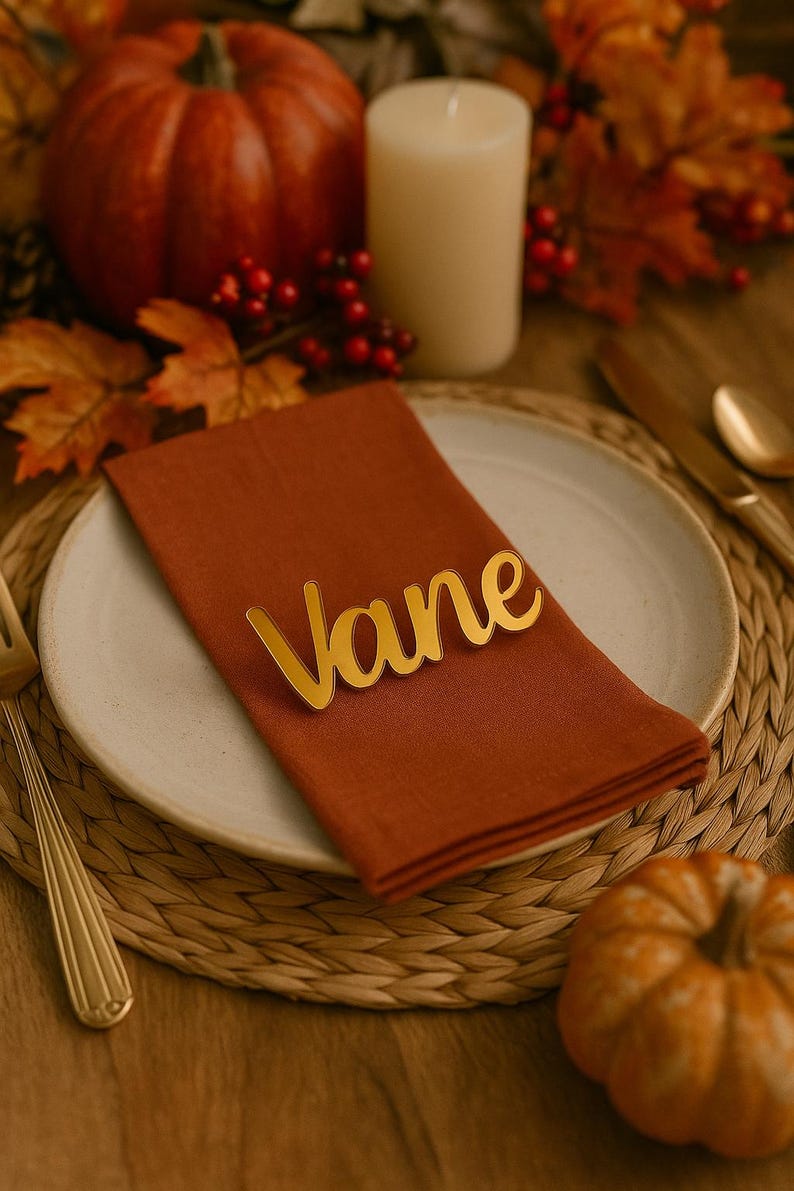 May include: A fall-themed table setting with a rust-colored napkin and a gold-colored nameplate that reads "Vane". The setting includes a white plate, a small pumpkin, and a candle. Autumn leaves and berries are also visible.
