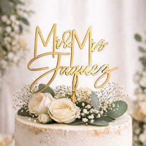 May include: A gold cake topper with the words "Mr & Mrs Jaquez" in a stylish font. The topper sits atop a white frosted cake adorned with white roses, baby's breath, and foliage. The cake is a wedding centerpiece.