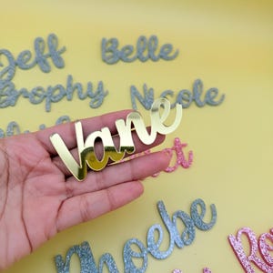 May include: A gold-colored acrylic name cutout that reads "Vane" is held in a hand. Other glittery name cutouts in silver and pink are scattered on a yellow background. The names are in a cursive font.