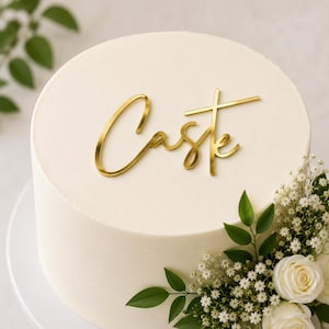 Puede incluir: Un pastel redondo de color blanco con un adorno de acrílico dorado que muestra la palabra Caste en letra cursiva. El pastel está decorado en un lateral con un conjunto de rosas blancas y pequeñas flores blancas con hojas verdes.