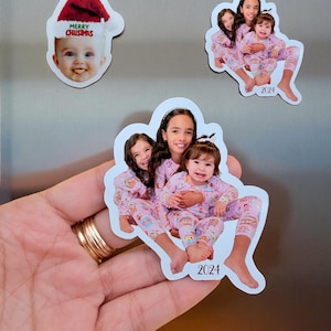 May include: Custom photo magnets featuring children in pyjamas and a baby in a Santa hat. One magnet has the text "Merry Christmas". The other magnets have the year "2024" printed on them. The magnets are white with a glossy finish.