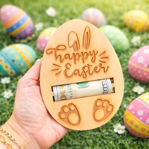 May include: A wooden Easter egg-shaped gift holder with the words "Happy Easter" engraved on it. The holder has a slot for money and bunny feet at the bottom. Colorful Easter eggs are in the background.