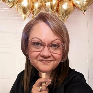 May include: A cardboard cutout of a woman with brown hair wearing pink-rimmed glasses. The cutout is held up by a stick.