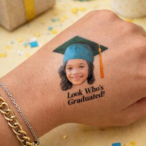 May include: A temporary tattoo of a smiling child wearing a graduation cap. The cap is teal with a gold tassel. The text "Look Who's Graduated!" is below the image. A gold and silver bracelet is on the wrist.