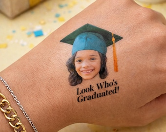 Peut inclure: Un tatouage temporaire présentant le portrait photographique d'un enfant souriant portant une toque de diplômé vert foncé avec un pompon orange. Sous le portrait, le texte noir indique, Look Who's Graduated! Le motif est appliqué sur la peau.