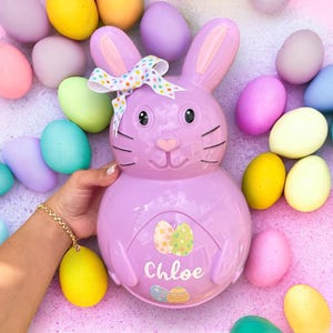 May include: A purple bunny-shaped Easter egg holder with a white bow and colourful polka dots. The holder has a cutout of two Easter eggs and the name "Chloe" written on it. The holder is surrounded by pastel-coloured Easter eggs.