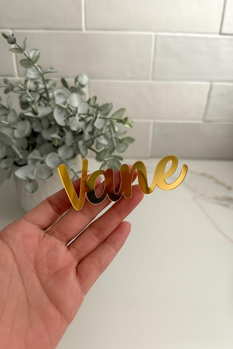 May include: A gold-colored acrylic name cutout that reads "Vane" in a cursive font. The name is held in a hand, with a blurred background of greenery and a white brick wall. The name is likely for a wedding or event.