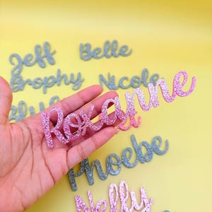 May include: A pink glittery "Roxanne" name cutout is held in a hand, with other glitter names in the background. The names are in a cursive font. The background is a solid yellow.
