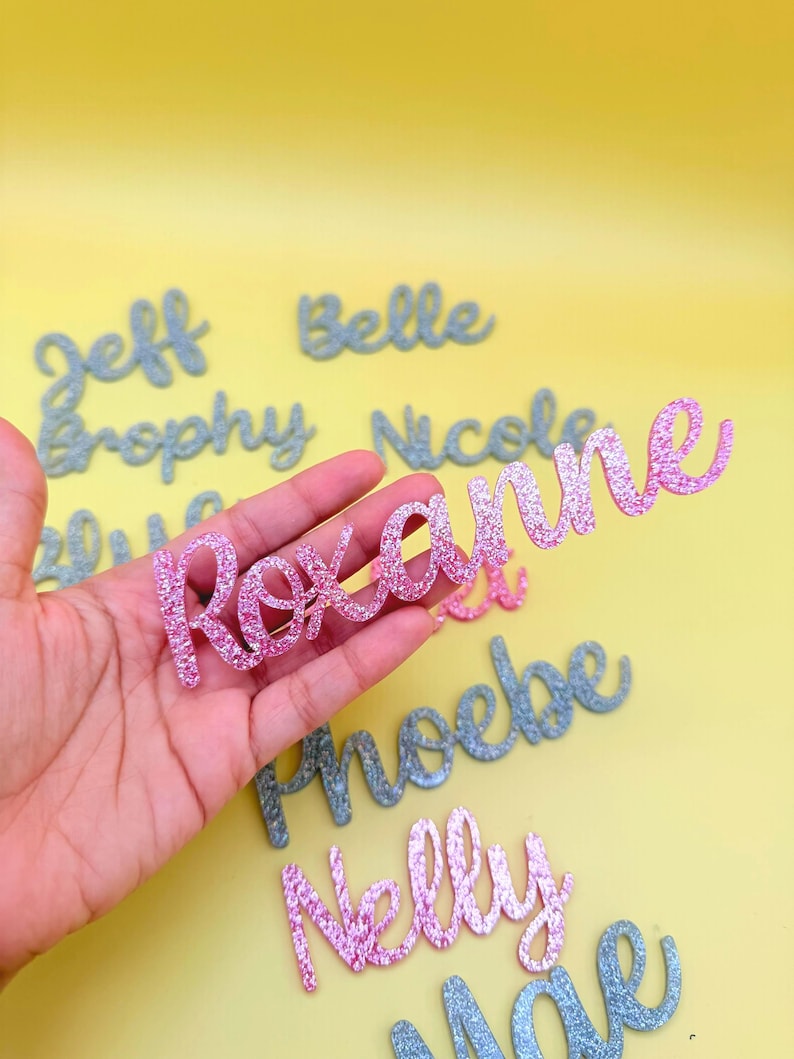 May include: A pink glittery "Roxanne" name cutout is held in a hand, with other glitter names in the background. The names are in a cursive font. The background is a solid yellow.