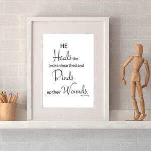 Peut inclure: Impression encadrée blanche avec une citation inspirante en noir et blanc qui dit "Il guérit les cœurs brisés et panse leurs blessures." La citation est tirée du Psaume 147:3. L'impression est sur une étagère blanche avec une figurine en bois, un cactus en pot et un appareil photo.