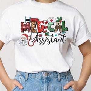 May include: White t-shirt featuring the words "MEDICAL ASSISTANT" in a festive, colourful design. The design incorporates medical symbols and a Santa Claus figure. The shirt is worn with blue jeans.