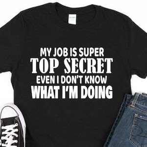 May include: Black t-shirt with the text "MY JOB IS SUPER TOP SECRET EVEN I DON'T KNOW WHAT I'M DOING" in white. The shirt is paired with blue jeans and black and white sneakers.