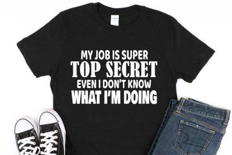 May include: Black t-shirt with the text "MY JOB IS SUPER TOP SECRET EVEN I DON'T KNOW WHAT I'M DOING" in white. The shirt is paired with blue jeans and black and white sneakers.