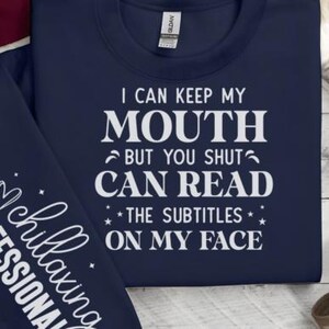 May include: Navy blue sweatshirt with white text that reads "I CAN KEEP MY MOUTH BUT YOU SHUT CAN READ THE SUBTITLES ON MY FACE." The sleeve has the text "chillaxing PROFESSIONAL."
