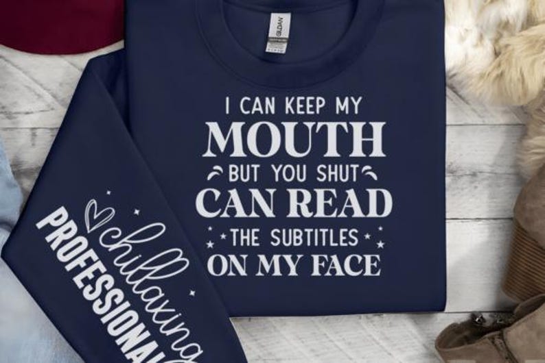 May include: Navy blue sweatshirt with white text that reads "I CAN KEEP MY MOUTH BUT YOU SHUT CAN READ THE SUBTITLES ON MY FACE." The sleeve has the text "chillaxing PROFESSIONAL."
