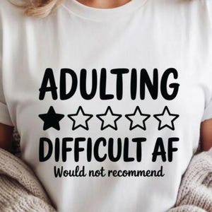 May include: White t-shirt with the text "ADULTING DIFFICULT AF Would not recommend" in black. The word "ADULTING" is above "DIFFICULT AF", with a black star and four white stars between them. The shirt is worn by a person.
