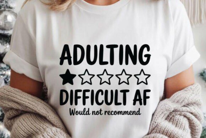 May include: White t-shirt with the text "ADULTING DIFFICULT AF Would not recommend" in black. The word "ADULTING" is above "DIFFICULT AF", with a black star and four white stars between them. The shirt is worn by a person.