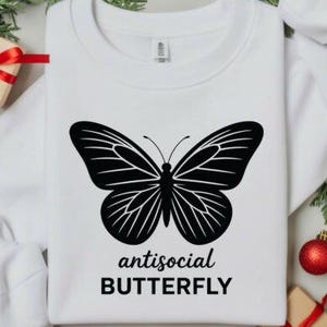 May include: White sweatshirt featuring a large black butterfly design with the words "antisocial BUTTERFLY" in a bold font. The design is centered on the front of the shirt. The sweatshirt is surrounded by holiday decorations.