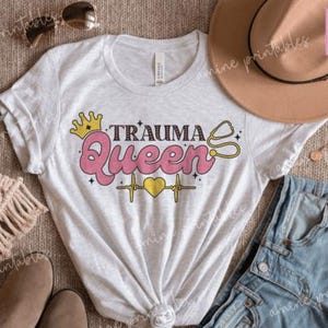 May include: A white t-shirt featuring the words "Trauma Queen" in pink, yellow, and black, with a crown, stethoscope, and heartbeat design. The image includes sunglasses, a hat, and denim shorts, styled on a neutral surface.