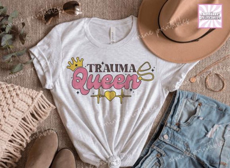 May include: A white t-shirt featuring the words "Trauma Queen" in pink, yellow, and black, with a crown, stethoscope, and heartbeat design. The image includes sunglasses, a hat, and denim shorts, styled on a neutral surface.