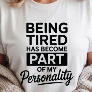 May include: White t-shirt with the black text "BEING TIRED HAS BECOME PART OF MY Personality". The shirt has short sleeves and is paired with a light-colored sweater.