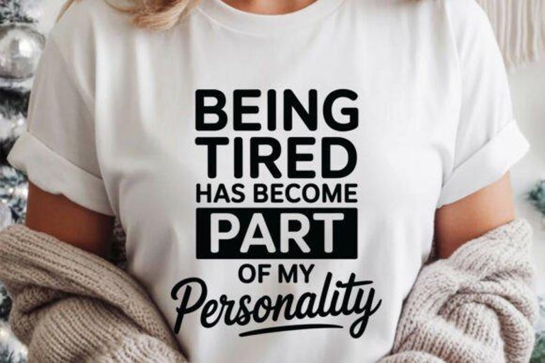 May include: White t-shirt with the black text "BEING TIRED HAS BECOME PART OF MY Personality". The shirt has short sleeves and is paired with a light-colored sweater.